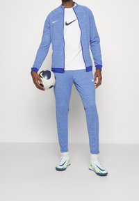 Blåt atletisk træningssæt lavet af tekstureret stof, med en lynlåstrøje og tilspidsede bukser. En hvid t-shirt under og en fodbold medfølger.