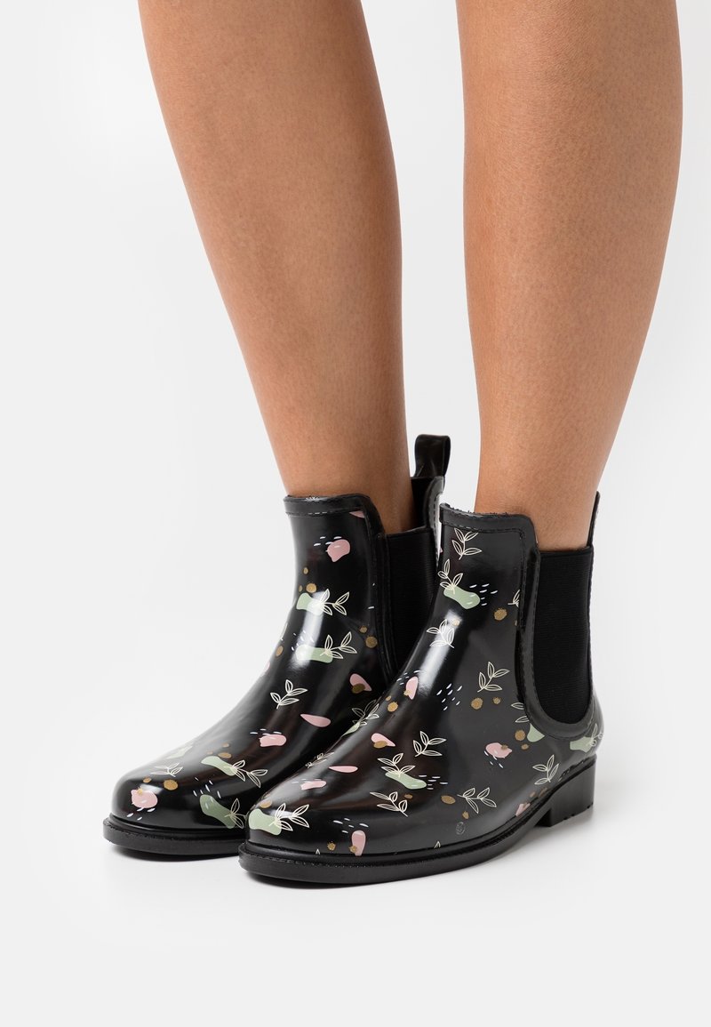 Bottes de pluie noires brillantes à la cheville avec des motifs floraux verts, roses et blancs portées sur des jambes nues sur fond blanc.