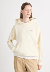 Hoodie de couleur crème avec une poche avant, arborant le texte brodé "Grant 1949" en marron. Matière douce, coupe décontractée et design à capuche.