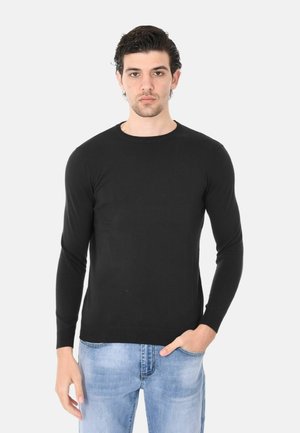 Giovane uomo con capelli scuri che indossa un maglione nero a maniche lunghe e jeans azzurri, in piedi con una mano in tasca.