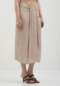 Jupe portefeuille beige en tissu léger, avec des plis à la taille ; assortie avec des sandales à talons strappy marron.