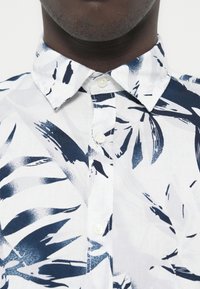 Κοντομάνικο πουκάμισο σε λευκό με σχέδια από φύλλα σε ναυτικό μπλε, με κολάρο που κλείνει με κουμπιά, μικρά λευκά κουμπιά και υφασμάτινη υφή.