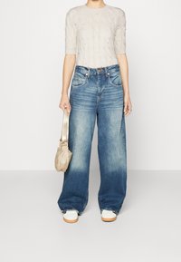 Osoba oblečená v krémovém pleteném krátkém svetru, širokých vybledlých modrých džínách, bílých teniskách a držící béžovou kabelku po svém boku.