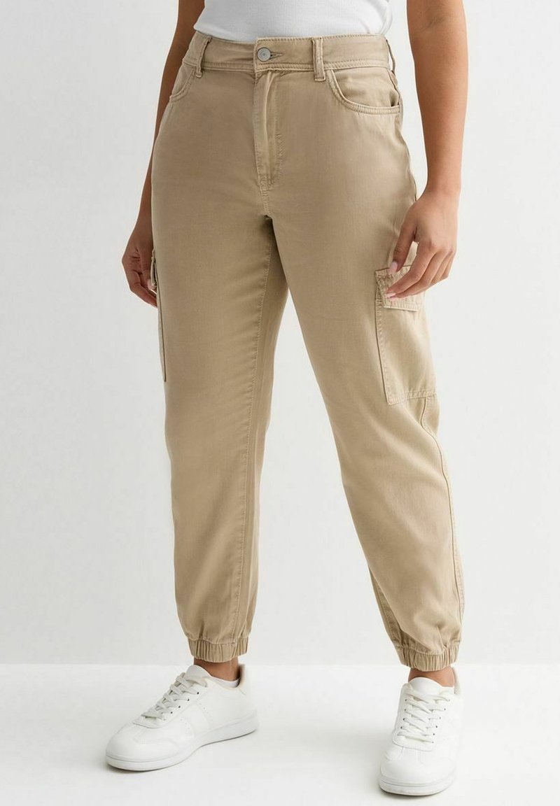 Personne portant un pantalon cargo beige avec des poignets élastiques et des baskets blanches, se tenant contre un fond clair et uni.