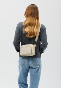 Borsa beige a tracolla realizzata in materiale liscio, con chiusura a zip, design rettangolare e tracolla regolabile, indossata sopra un maglione grigio.