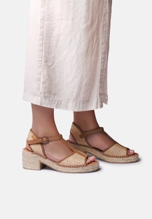 Feet wearing tan woven T-strap block heel sandals and cropped white linen pants against a plain light background.