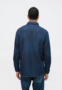 Σκούρο μπλε τζιν πουκάμισο με κολλάρο που κουμπώνει, μακρύ μανίκι και άνετη εφαρμογή. Διαθέτει ευθύ τελείωμα και διακριτές ραφές.