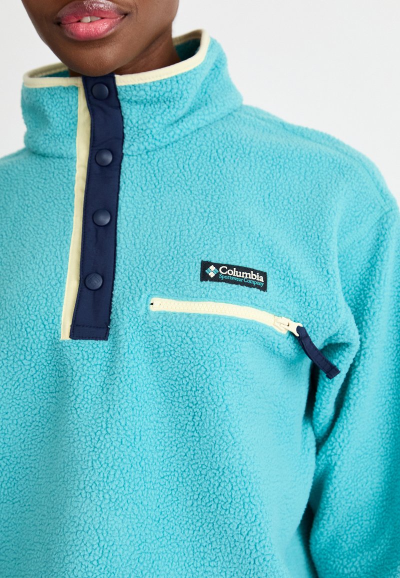 Persona vestida con un pulóver de forro polar turquesa de Columbia, con un alto collar, botones a presión en color navy y un bolsillo en el pecho con cremallera de color crema.