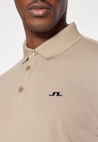 Bēša polo krekls ar trīs pogu pogām, īsām piedurknēm un melnu logo uz krūtīm. Izgatavots no gludas, struktūrētas auduma.