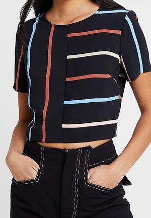 Kvinde iført en sort cropped top med farverige lodrette og vandrette striber, kombineret med sorte bukser med synlige hvide syninger.