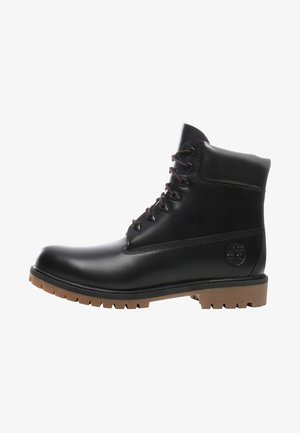 Bottine en cuir noir avec des lacets marron, bout rond et semelle en caoutchouc texturé. Elle présente un logo en relief sur le côté extérieur.