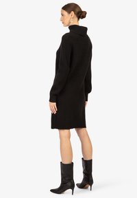 Schwarzes Strickkleid mit hohem Kragen, langen Ärmeln und lockerer Passform, versehen mit gerippter Textur und seitlichen Schlitzen, kombiniert mit schwarzen Stiefeln mit High Heels.