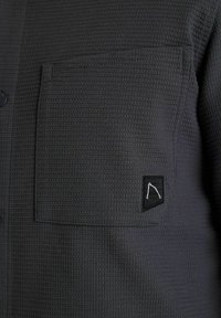 Dunkelgraues strukturiertes Hemd mit einer aufgesetzten Tasche, die ein schwarzes Etikett mit einem minimalistischen weißen Logo zeigt. Funktionale Knöpfe entlang der Vorderseite.