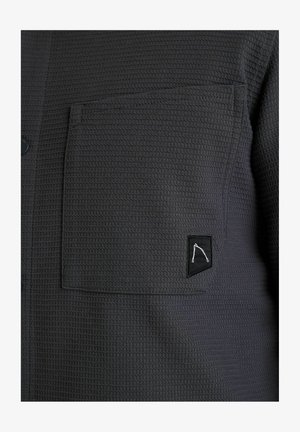 Tmavosivá textúrovaná košeľa s náprsní vreckom, na ktorom je čierna etiketa s minimalistickým bielym logom. Funkčné gombíky po celej prednej strane.