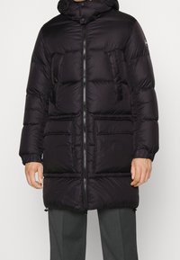 Svart pufferjacka med huva, dragkedja fram och flera framfickor; slät textur med kviltade sektioner för isolering.