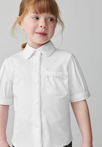 Kortärmad vit skjorta med krage, knappar ned framtill och en liten ficka med en rosett. Slät tygstruktur.