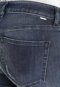 Jeans de mezclilla oscura que presentan un diseño clásico de cinco bolsillos con costuras visibles y una pequeña etiqueta de logo plateado en el bolsillo trasero.