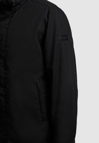 Black long-sleeve jacket with front zipper and side pocket, featuring a small "KHUJO" logo patch on the upper arm.