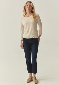 Bézs színű kötött pulóver kerek nyakkivágással, háromnegyedes ujjakkal, sötétkék, testhez álló farmerrel és világosbarna velúr sneakerrel párosítva.