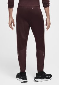 Burgundowe spodnie sportowe o tapered designie, z panelem dekoracyjnym, odblaskowymi akcentami i elastycznymi mankietami, w połączeniu z czarnymi butami sportowymi.