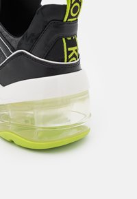Talons de baskets noirs avec languette en tissu vert néon, semelle intermédiaire blanche et semelle amortie transparente à coussin d'air sur semelle extérieure vert néon sur fond blanc.