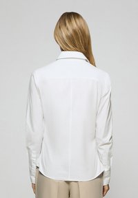 Vit långärmad skjorta med spetsig krage, slät textur, knäppta manschetter och rundad nederkant, sedd bakifrån.