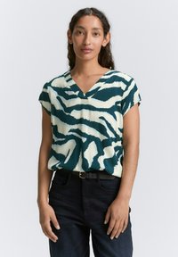 Blouse courte à manches courtes vert et crème avec décolleté en V et motif abstrait de zèbre. Portée avec un pantalon en denim foncé. Texture lisse.