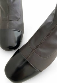 Botas de tobillo de cuero marrón con punteras de charol negro, que presentan costuras visibles y una textura suave.