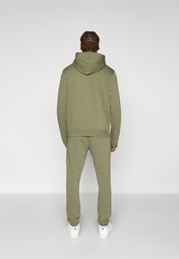 Olīvzaļš kapuces krekls un saskaņotas sporta bikses, kas izgatavotas no mīkstas kokvilnas maisījuma. Ievērojamas iezīmes ir kapuce, ribotas aproces un potīšu aproces. Balti sporta apavi papildina izskatu.