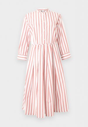 Kobieta stoi z rękami na biodrach, ubrana w białą sukienkę midi z pomarańczowymi pionowymi paskami i czarne japonki na jednolitym tle.