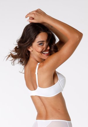 Donna con capelli castani che sorride, indossando un reggiseno bianco e biancheria intima trasparente bianca, posando con le braccia alzate davanti a uno sfondo neutro.