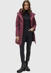 Maroon waterproof coat with a hood, zip and snap closures, drawstring waist, and side pockets, paired with black jeans and ankle boots.