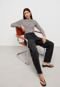 Randig långärmad topp i svart och vitt, kombinerad med höga svarta jeans och svarta flip-flops, sittande på en stol med kromram och orange sits.