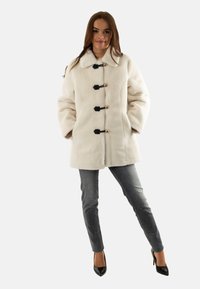 Cappotto in pelliccia sintetica bianca con ampio colletto, bottoni a gancio frontali neri e dorati, abbinato a jeans skinny scuri e tacchi neri.