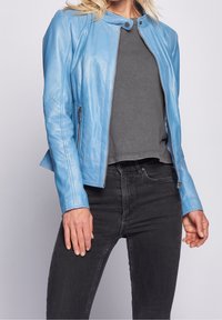 Kvinde iført lysblå læderjakke over grå skjorte og sorte jeans, poserer foran en ensfarvet lys baggrund.