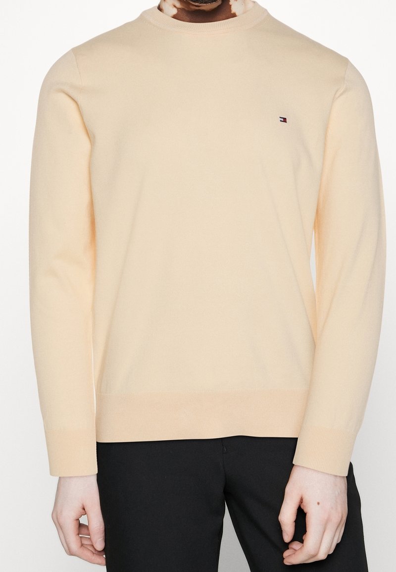 Homme portant un pull beige clair à manches longues avec un petit logo Tommy Hilfiger sur la poitrine et un pantalon noir.