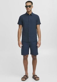 Conjunto de camisa de manga corta y pantalones cortos de color navy, confeccionado en tejido ligero, acompañado de gafas de sol oscuras y sandalias marrones, de pie sobre un fondo neutro.