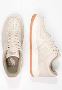 Par av beiga Nike Air Force 1 sneakers med texturerat mönster och orange gummisulor, en med ovanvy, den andra med sidovy.