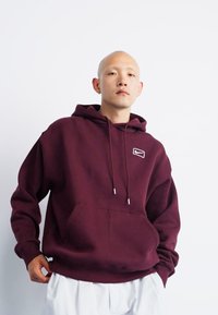 Maroon hoodie with a drawstring, front pocket, and a small logo. Made from a soft fabric with ribbed cuffs and hem. Casual design.