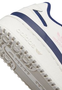 Bijele tenisice s mornarsko plavim akcentima, teksturirana gumena potplat s kružnim uzorcima, te "adidas" logo urezan sa strane i na peti.