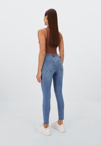 Jeans ajustados azules de cintura alta, con dos bolsillos traseros y una textura suave. Combinados con zapatillas blancas y una blusa sin mangas marrón.
