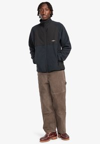 Fleece jacket in navy and black, with a zip front, high collar, and side pockets. Paired with brown, textured corduroy pants and brown shoes.