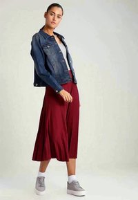 Woman wearing a dark blue denim jacket, white top, red midi skirt, white socks, and gray sneakers against a plain light background.