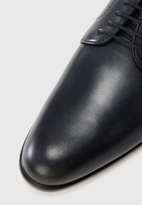 Chaussure habillée en cuir noir avec un bout pointu, une texture lisse et un design à lacets. Œillets métalliques remarquables et éclat subtil.