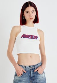 Hvit ribbestrikket crop topp med "RACER" i rødt og blått. Modellen har på seg lyseblå, slitte denimshorts, med hendene i lommene, og står mot en hvit bakgrunn.