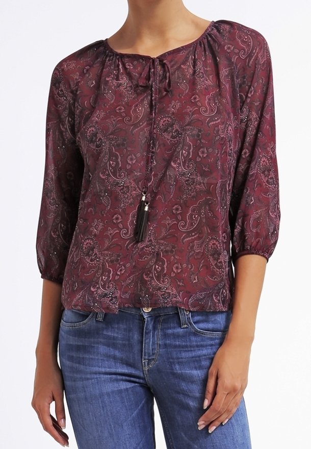 Femme portant un chemisier bordeaux à motif cachemire avec des manches trois-quarts et un col à nouer avec pompons, assorti à un jean bleu.