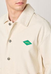 Beige Canvasjacke mit spitzem Kragen, ausgestattet mit silbernen Knopfverschlüssen und einem grünen, rautenförmigen Logoaufnäher auf der linken Brust.