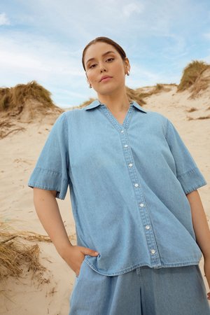 Femme debout sur des dunes de sable portant une chemise en denim bleu clair ample et un short assorti, une main dans la poche.