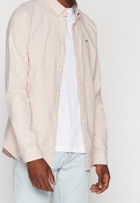 Man in een beige overhemd over een wit T-shirt en lichtblauwe jeans, staande tegen een effen lichte achtergrond.