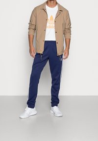 Béžová lehká bunda přes bílý tričko s oranžovým logem, v kombinaci s námořnickými tepláky se bílými pruhy a bílými sportovními botami.